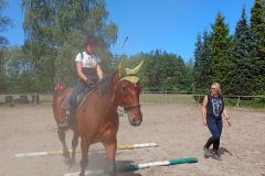 Uczestniczka wakacyjnej szkółki jeździeckiej pokonuje na koniu przeszkodę z leżących  drągów pod czujnym okiem instruktorki